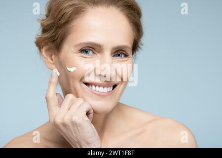 Donna di mezza età che applica crema e cosmetici anti-età sul viso, su sfondo blu. Cura della pelle per le donne con cosmetici anti-età Foto Stock