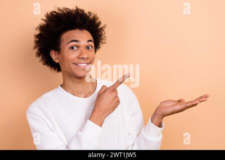 Foto di un giovane sorridente indossa abiti bianchi alla moda e presenta il nuovo logo prodotto del marchio spazio vuoto isolato su sfondo di colore beige Foto Stock