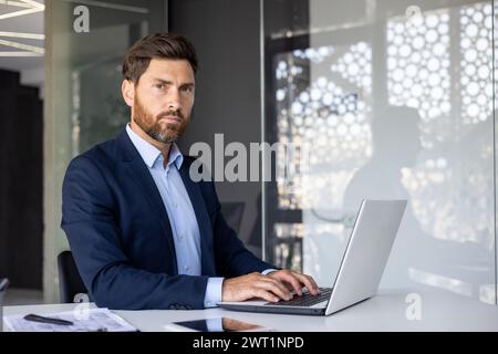 Ritratto di un giovane uomo d'affari serio e sicuro di sé seduto in ufficio al tavolo, scrivendo su un computer portatile, lavorando con clienti e documenti, guardando la fotocamera. Foto Stock