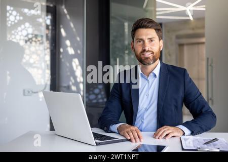 Ritratto di un giovane avvocato e politico uomo sorridente e di successo seduto in ufficio al tavolo con un portatile e guardando, con orgoglio, la macchina fotografica. Foto Stock