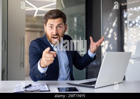 Giovane uomo d'affari arrabbiato seduto in ufficio al tavolo e puntando il dito verso la telecamera, abituandosi al problema e all'errore, urlando scontento. Foto Stock
