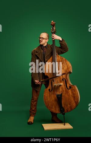 Uomo con baffi eleganti in tuta marrone che suona il contrabbasso su sfondo verde. Evento di musica classica Foto Stock