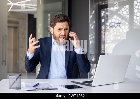 Giovane uomo d'affari, arrabbiato, seduto a una scrivania in ufficio e che parla aggressivamente al telefono, grida e gesti con le mani. Foto Stock