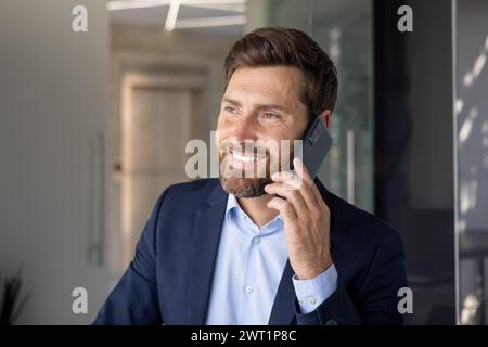 Ritratto di un giovane uomo d'affari sorridente felice, avvocato che è in ufficio e parla al telefono. Guarda di lato, una foto da vicino. Foto Stock