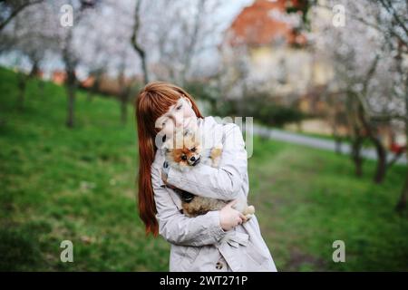 Una ragazza dai capelli rossi tiene un cane Spitz rosso tra le braccia e cammina nel giardino di primavera Foto Stock