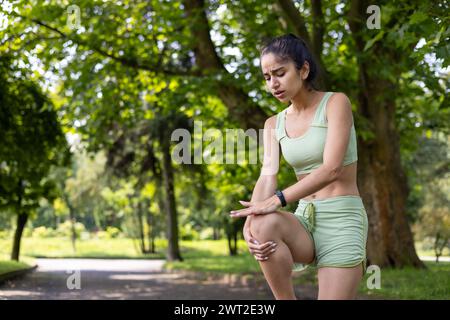 La giovane ragazza indiana in piedi in un parco con una tuta e tenendo le mani sul ginocchio, sentendo molto dolore, si spruzzò i muscoli dopo aver corso e fatto sport. Foto Stock