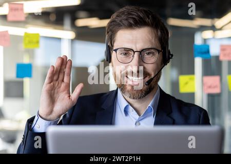 Uomo d'affari professionale che indossa una cuffia e sorride durante una videoconferenza in un ufficio moderno. Foto Stock