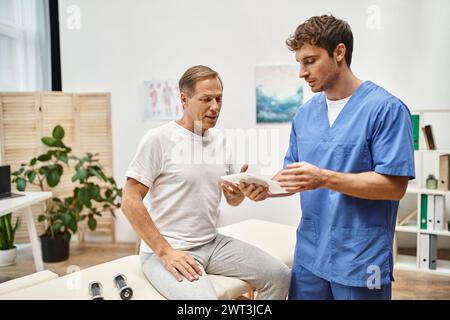 bel medico in abito blu che tiene la compressa e ascolta il suo paziente maturo durante l'appuntamento Foto Stock