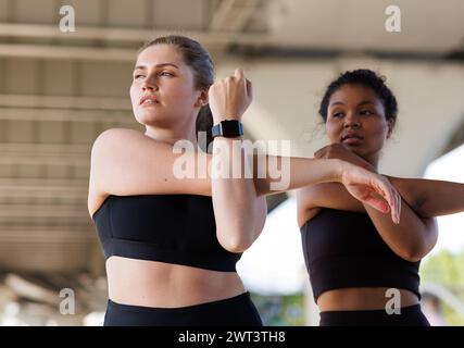 Due femmine allungano le mani mentre si esercitano sotto un ponte. Giovane donna di grandi dimensioni che fa esercizi di riscaldamento con la sua amica e guarda lontano. Foto Stock