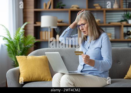 Una donna matura che sembra sorpresa e ansiosa mentre tiene in mano una carta di credito e utilizza un laptop sul divano di casa. Foto Stock