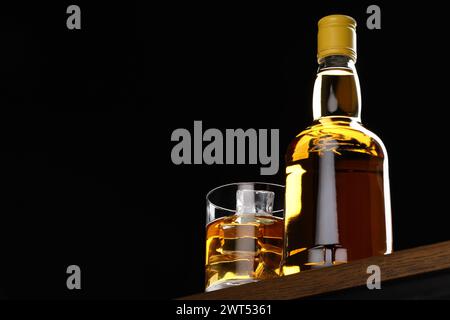 Whisky con cubetti di ghiaccio in vetro e bottiglia su tavolo in legno su sfondo nero, vista ad angolo basso. Spazio per il testo Foto Stock