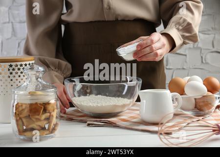 Donna che aggiunge lievito in polvere nel recipiente sul tavolo di legno bianco, primo piano Foto Stock