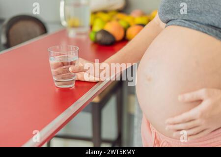 Abbracciando i benefici vitali dell'acqua durante la gravidanza, una donna incinta sta in cucina con un bicchiere, evidenziando il ruolo cruciale dell'idratazione in Foto Stock