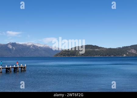 Foto orizzontale di un enorme lago calmo con un molo sul lato, e montagne piene di vegetazione poste in lontananza coperte da nebbia, e un Foto Stock