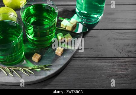 Absinthe in bicchieri, rosmarino, zucchero di canna e lime su un tavolo in legno nero, spazio per il testo. Bevanda alcolica Foto Stock