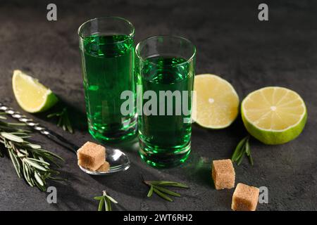 Absinthe in bicchieri da shot, cucchiaio, zucchero di canna, lime e rosmarino su un tavolo testurizzato grigio, primo piano. Bevanda alcolica Foto Stock