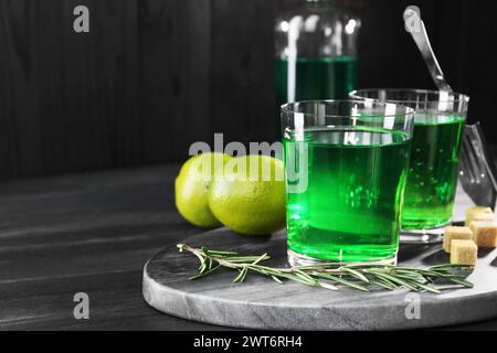 Absinthe in bicchieri, rosmarino, zucchero di canna e lime su un tavolo in legno nero, spazio per il testo. Bevanda alcolica Foto Stock