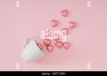 Composizione d'amore creativa realizzata con cuori di cioccolato avvolti in lamina rosa che escono dalla tazza da caffè bianca su sfondo rosa pastello. Concetto d'amore minimalista. Foto Stock