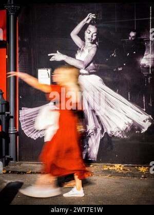 Una donna passa davanti a un poster di balletto a grandezza naturale, la sua figura assomiglia a quella della ballerina che offre un'interessante fotografia di strada giustapposta a Londra Foto Stock