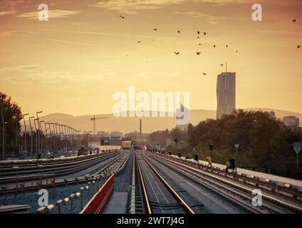 Linea della metropolitana sopraelevata in direzione di Donaucity, con una vista prospettica in diminuzione. Tramonto in città, montagne all'orizzonte. Stormo di uccelli che volano sopra. Foto Stock