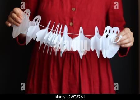 Una donna in abito rosso tiene in mano la carta bianca fantasma garland. Foto ravvicinata delle decorazioni di Halloween Foto Stock