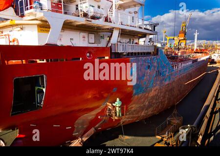 Il lavoratore dipinge di rosso lo scafo della nave da carico nel bacino di carenaggio sotto il cielo blu. Manutenzione di navi marittime, lavori di verniciatura industriale nei cantieri navali. Grande autocisterna Foto Stock