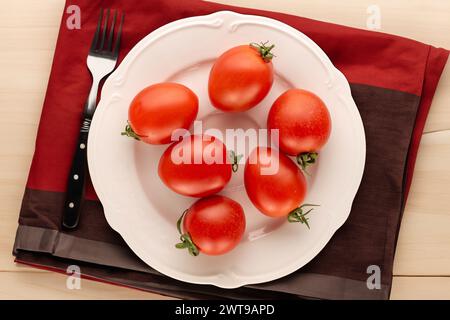 Diversi pomodori da cocktail su un piatto di ceramica bianca con un tovagliolo e una forchetta su un tavolo di legno, vista dall'alto. Foto Stock