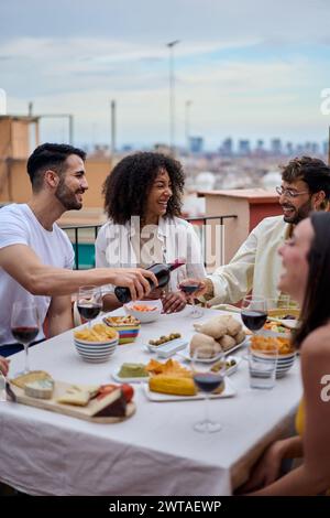 Giovane uomo caucasico sorridente che serve vino rosso agli ospiti al tavolo per festeggiare gli amici felici. Foto Stock