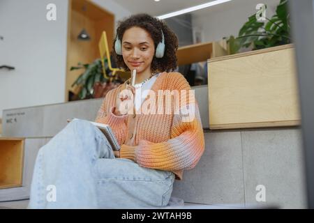 Una donna con le cuffie si siede sulle scale di una stanza, tenendo in mano una penna e sorridendo Foto Stock