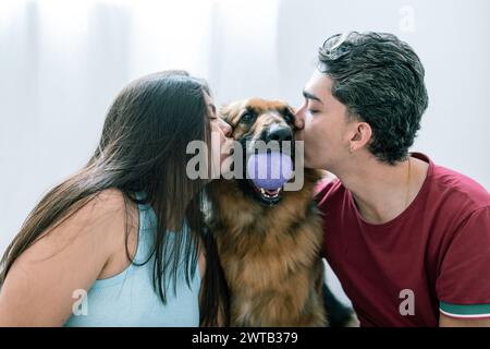 Felice coppia latinoamericana che condivide un bacio con il loro sorridente cane da pastore tedesco Foto Stock