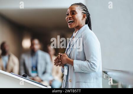 Donna africana professionale con un sorriso radioso coinvolge i partecipanti mentre parla a una conferenza d'affari. Con una squadra sfocata sullo sfondo, emb Foto Stock