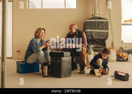 Durante un progetto di ristrutturazione di una casa, una famiglia si prende una pausa. Mamma e papà seduti insieme, bevono un caffè mentre il figlio gioca nelle vicinanze. La famiglia mi sta mettendo Foto Stock