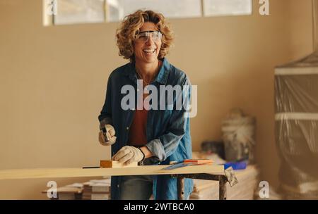 Una donna caucasica felice ristruttura la sua casa, lavorando su basi in cucina. Inchioda abilmente un pannello di legno con un martello, utensili manuali e sicurezza Foto Stock