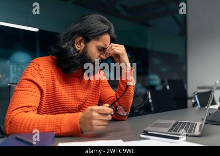 Esausto e sovraccarico di lavoro, giovane indiano all'ufficio buio Foto Stock