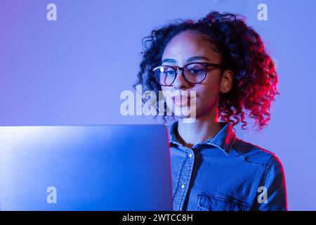 Donna nera concentrata con occhiali che lavorano su un notebook con illuminazione blu Foto Stock