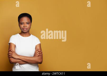 Donna afroamericana seria e sicura di sé con t-shirt bianca in piedi su sfondo giallo colorato Foto Stock