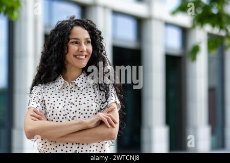 Donna professionista con una camicia a pois, in piedi con le braccia incrociate all'esterno di un moderno edificio per uffici. Foto Stock