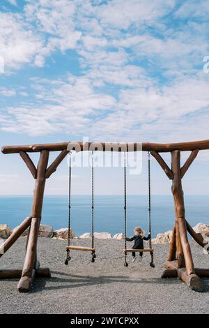 La bambina si siede su una grande altalena di legno sulla riva del mare e guarda lateralmente. Vista posteriore Foto Stock