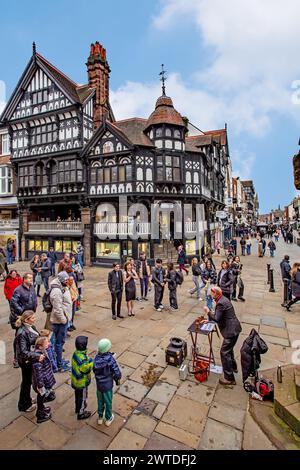 Gli amanti dello shopping camminano su marciapiedi bagnati e ciottoli nella città romana di Chester, in Inghilterra, intorno alla croce medievale e ai negozi nelle file Foto Stock