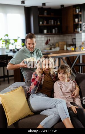 Una famiglia di tre persone si trova comodamente adagiata su un divano, con i volti che riflettono l'emozione e l'attenzione mentre condividono una ciotola di popcorn durante una sosp Foto Stock