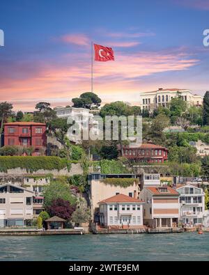 Foto al tramonto di una fila di case tradizionali su una collina verde sul Bosforo a Istanbul, Turchia, con una grande bandiera turca che sventola in alto. Gli alberi in primo piano sono lussureggianti e verdi Foto Stock