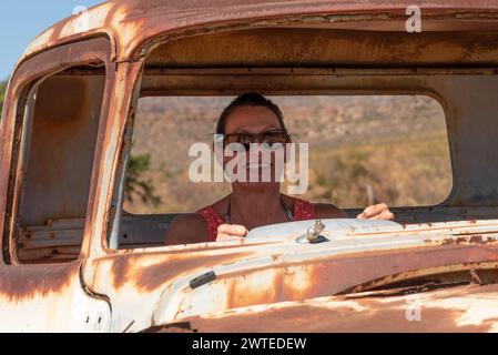 Sudafrica. 16/02/2024. Donna attraente che posa con una vecchia auto americana arrugginita sul ciglio della strada nella regione Swartland del Sudafrica. Foto Stock