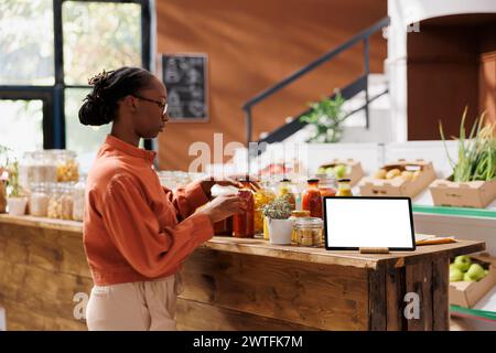 Cliente afroamericana che naviga nel punto vendita vicino a un tablet che mostra un modello di cromakey isolato. Donna nera in piedi accanto a un dispositivo digitale con uno schermo bianco vuoto. Foto Stock