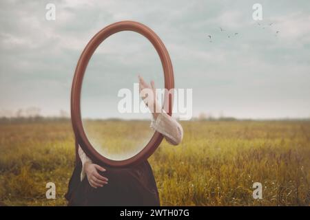 la donna si nasconde tenendo uno specchio davanti al viso e lo accarezza dolcemente con la mano; concetto di auto-amore Foto Stock