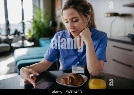 Infermiera o medico si prepara al lavoro la mattina, scorrendo sullo smartphone mentre fa colazione. Equilibrio vita-lavoro per gli operatori sanitari. Foto Stock