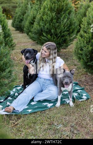 Bionda (33) ragazza abbraccia e sorride ai suoi cani di salvataggio dei box in fattoria di alberi Foto Stock