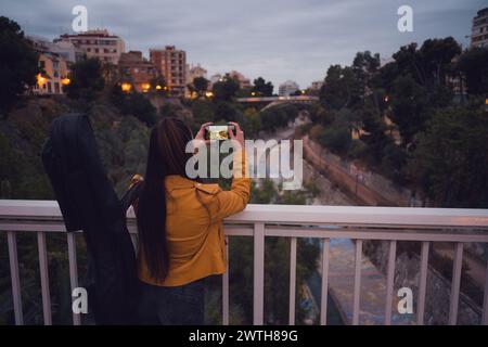 Donna con una chitarra che scatta foto del fiume dal ponte urbano al crepuscolo Foto Stock