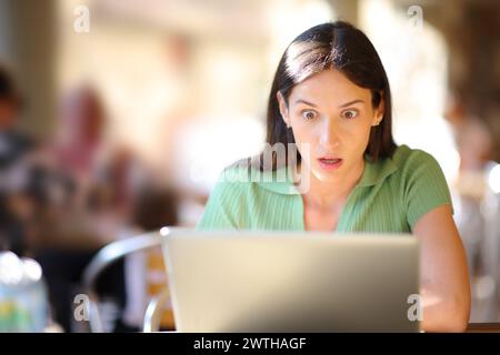 Vista frontale ritratto di una donna stupita che guarda i contenuti del notebook in una terrazza del bar Foto Stock
