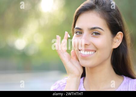 Ritratto di una donna felice che applica idratante che ti guarda in un parco Foto Stock