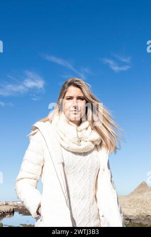 Una donna che indossa una giacca bianca e una sciarpa si trova di fronte a un cielo blu. Sembra sicura di sé e pronta, con i capelli che soffiano nel vento Foto Stock